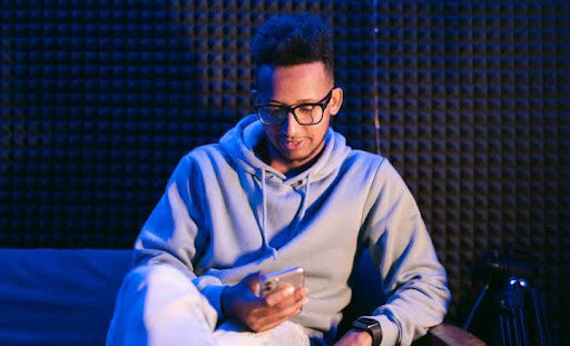 Can you wear blue light glasses all day: Close-up portrait of an attractive curly guy sitting on sofa using smartphone browsing web service at modern loft industrial style interior splashed by blue light
